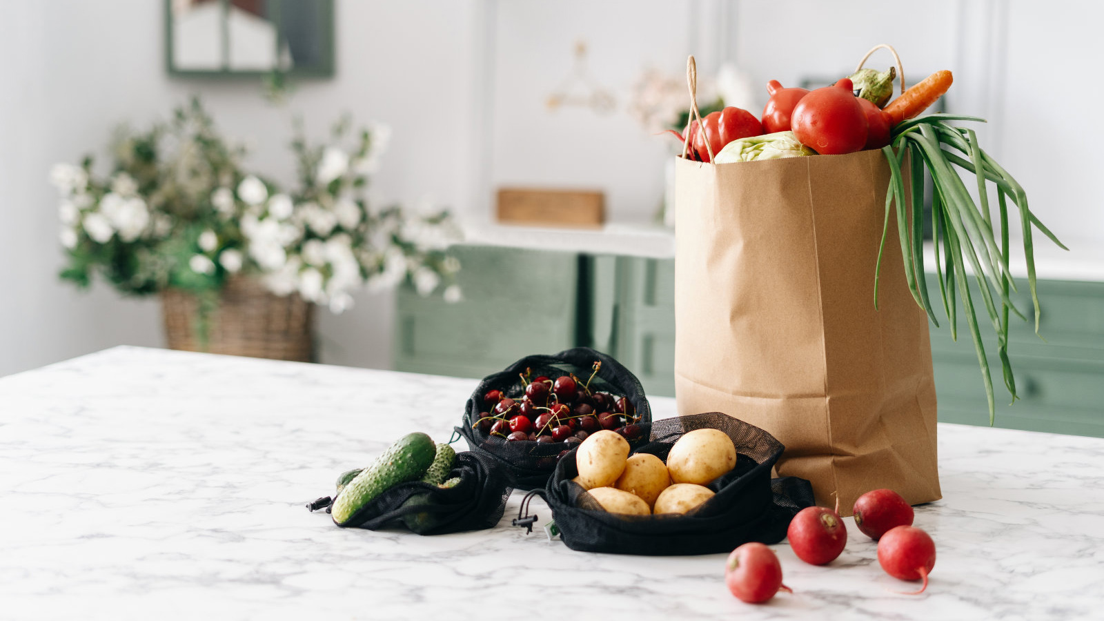 Various fresh vegetables in paper grocery and black mesh bags on kitchen island with marble top, healthy food full of nutrients, vintage furniture with flowers, blurred background
